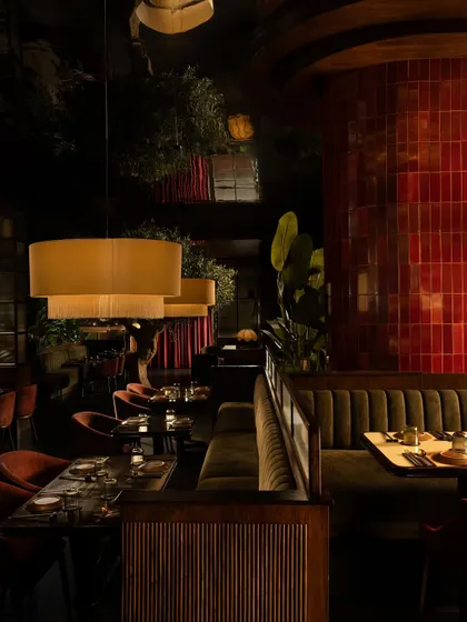 The dining area at Hikki, where a column of glossy red tiles punctuates the space. The mirrored ceiling reflects the warm lighting and lush greenery, creating an infinite, dreamlike quality in this intimate restaurant interior.