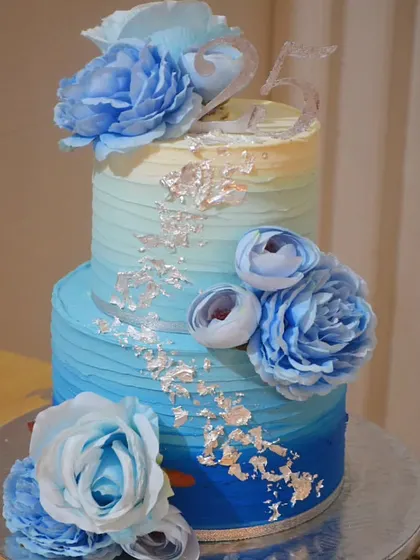 A beautiful two-tier anniversary cake in shades of blue. The ombre buttercream and silver leaf accents are complemented by stunning blue sugar flowers.