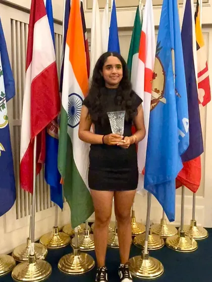Mahreen Bhatia proudly holds her trophy after finishing 4th at the US Kids World Teen Championships. She stands before a backdrop of international flags, highlighting her achievement on a global platform.
