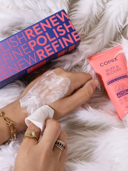 This lifestyle shot shows the texture of a face scrub on a model's hand against a soft, fluffy background. It highlights the gentle exfoliating action and luxurious feel of the product.