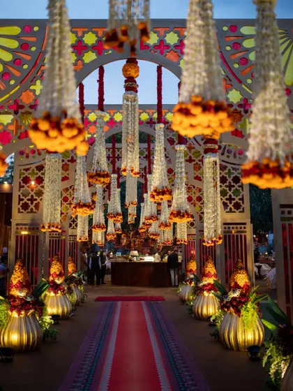 A grand entrance for a traditional event, lined with ornate floral 'Kalash' arrangements and featuring a walkway covered in hanging jasmine and marigold chandeliers. The design is opulent and deeply symbolic.