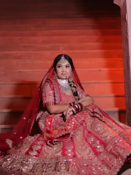 A full-length shot of a bride in a red lehenga, seated on grand steps against a warm, red-lit background. This pose highlights the volume and intricate work of the bridal skirt.
