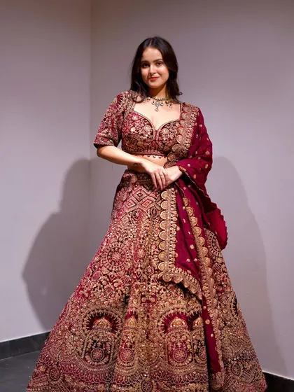 A full-length shot of a bride in a deep maroon velvet lehenga. The heavy gold embroidery gives it a very royal and luxurious feel, ideal for a grand wedding celebration.