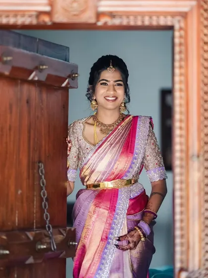 A full view of the bride in her pink and lavender saree, standing in a traditional doorway. The intricate blouse and rich saree colors make for a stunning bridal portrait.