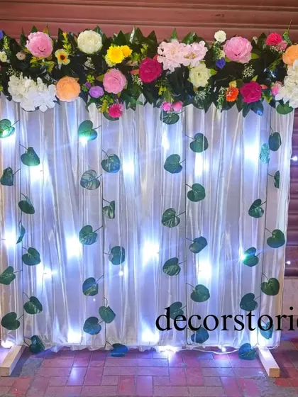 A nature-inspired ready-to-install backdrop for Ganpati. This design features cool white lights behind a sheer curtain, decorated with green leaf creepers and a colorful floral border at the top.