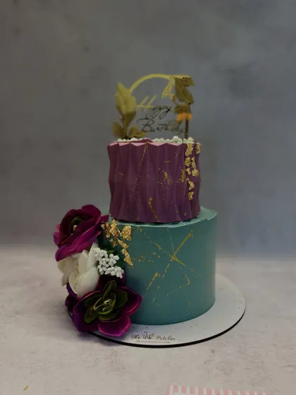 This two-tiered cake features a unique color combination of teal and purple. The top tier has a geometric texture, and the cake is finished with gold splatters and a cascade of sugar flowers.