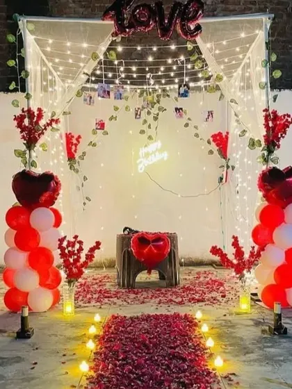 An outdoor romantic setup for a birthday or anniversary. It features a canopy with fairy lights, a rose petal aisle, and red and white balloon pillars.