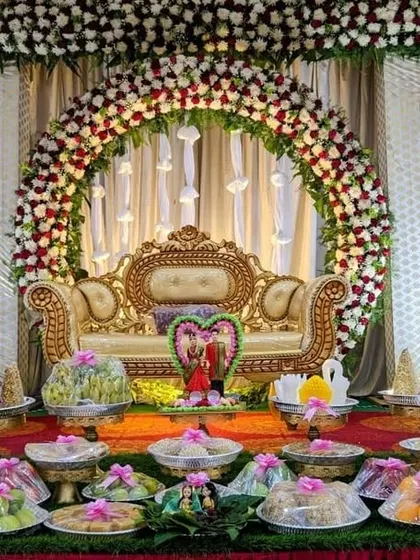 A grand engagement stage setup. It features a large floral arch, an ornate golden sofa, and a full display of decorated offering plates, blending tradition with elegance.