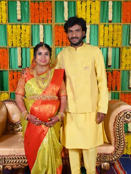A happy couple at their Seemantha ceremony. The backdrop is a vibrant wall of fresh flowers in a checkerboard pattern, creating a colorful and joyous setting.