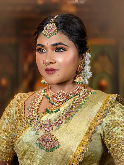 A close-up portrait of a bride in a golden-yellow saree. The focus is on her expressive eyes, defined by kohl, and the multi-layered traditional jewelry.