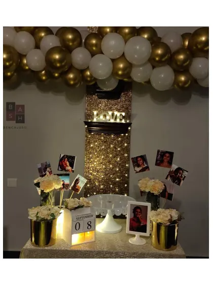 We can transform even a simple wall into a stunning feature. This date night setup includes a shimmering curtain of fairy lights, a personalized photo display table, and a beautiful gold and white balloon garland.