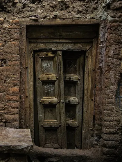 Tucked into a mud-brick wall, this dark, intricately carved wooden door is slightly ajar. The deep shadows and earthy tones give it a timeless and almost secretive quality.