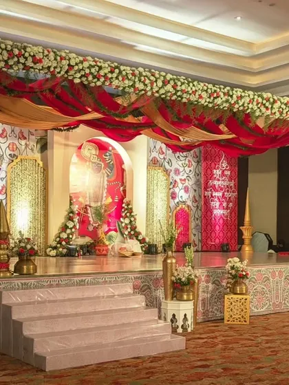 This stage is a beautiful example of traditional Indian wedding decor. It features a backdrop with classic motifs, draped in red and gold fabrics, and a wooden mandap structure adorned with florals, creating a rich, cultural ambiance.