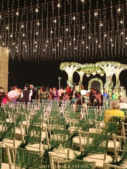 An evening view of a spectacular wedding, with guests seated on green-draped chairs under a sky of fairy lights, all focused on the beautifully lit floral stage.