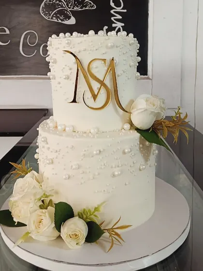 This sophisticated two-tiered wedding cake is decorated with edible pearls and a custom gold acrylic "MS" monogram. Fresh white roses and gold leaves complete this elegant design.