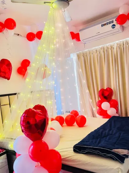 A romantic room setup with a red and white theme. A beautiful canopy made of net and fairy lights hangs over the bed, surrounded by red and white balloons and heart-shaped foil balloons, creating a dreamy ambiance.