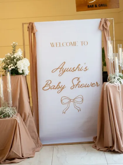 A classic and elegant welcome sign for Ayushi's baby shower, featuring beautiful typography and set against a backdrop of soft, mocha-colored drapes and white floral arrangements.