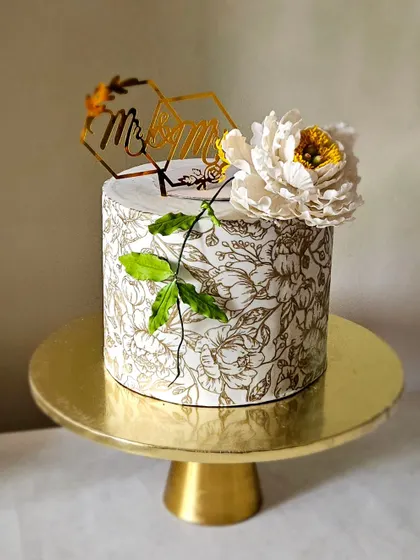 A chic and modern anniversary cake. This design features a delicate floral mesh stencil in gold on a white fondant base, topped with a large, handcrafted white sugar peony.