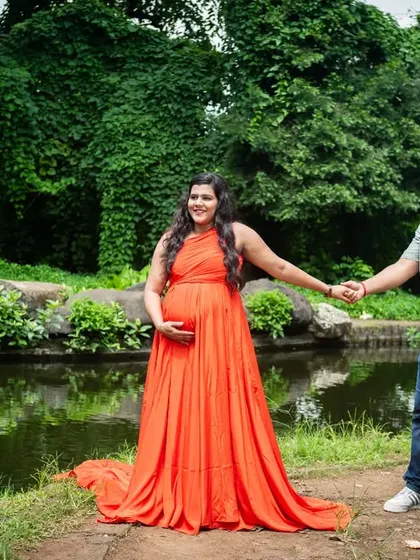 The vibrant orange gown stands out beautifully against the lush green backdrop of the park. This wide shot captures the full elegance of the dress and the happy, expectant energy of the mother-to-be.