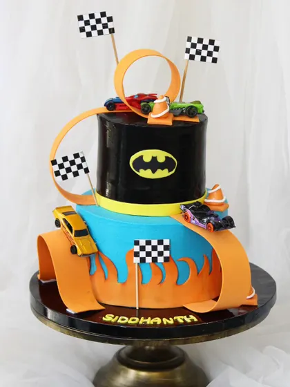 A dream cake for a Hot Wheels fan. This two-tiered design features a winding orange track with real Hot Wheels cars racing around a Batman-themed tier, complete with checkered flags for a thrilling finish.