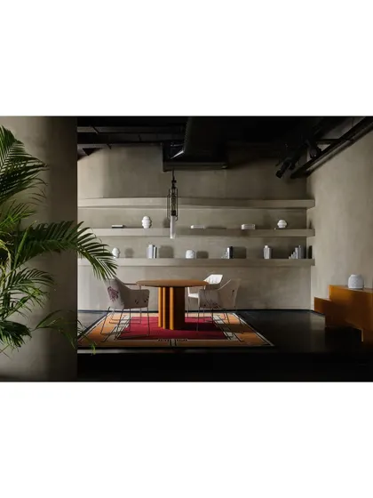 A wider view of the dining setup, showing how the colorful rug anchors the space. The design uses a mix of materials, from the metal legs of the chairs to the wooden table and concrete-finish walls.