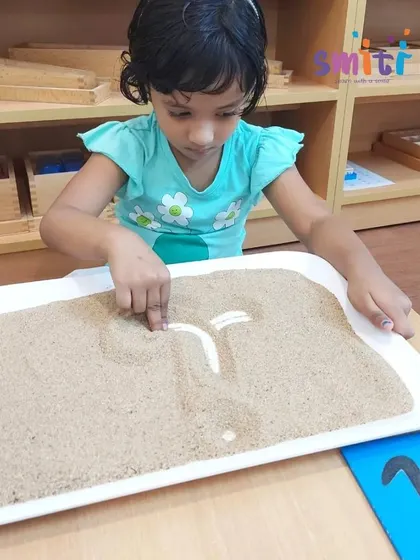 A young girl traces a cursive letter in a sand tray, a tactile way to build muscle memory for writing. This pre-writing exercise is a key step in our language curriculum.