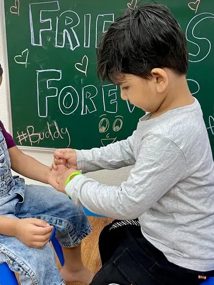 A child carefully ties a friendship band on his friend's wrist. These moments help instill the values of sharing, caring, and celebrating one another.