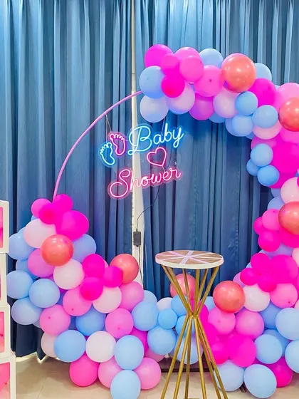 A vibrant baby shower setup with a balloon ring in pink and blue. The decoration includes a custom "Baby Shower" neon sign and "BABY" blocks.