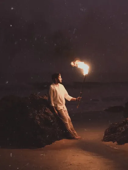 A solo portrait of the groom-to-be, sitting contemplatively on a rock by the sea while holding a lit torch. The image has a grainy, film-like quality, adding to the moody and romantic atmosphere of the Goa pre-wedding shoot.