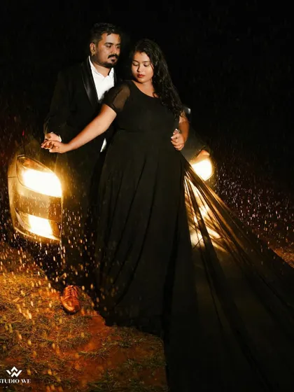 A cinematic shot of a couple standing in front of car headlights in the rain. The dramatic lighting and flowing dress create a scene straight out of a movie.