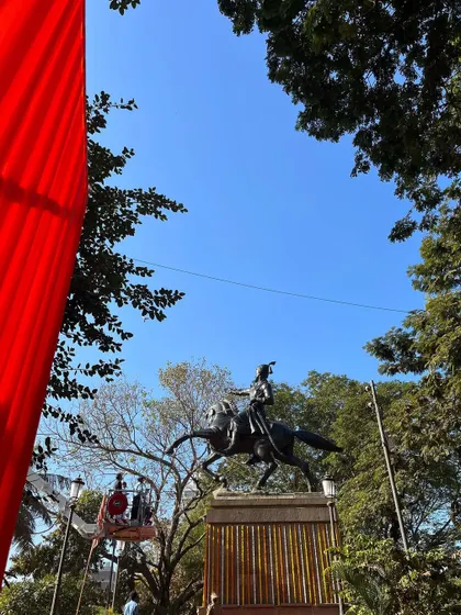 A wide shot of the Shivaji Maharaj statue at Shivaji Park, with a large red flag in the foreground during Jayanti celebrations.