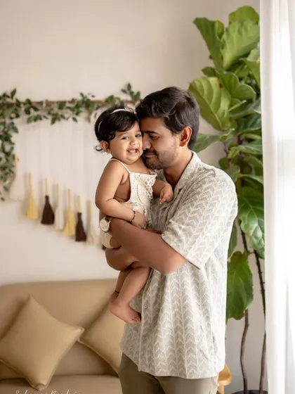 A father holds his daughter, and they share a sweet, smiling moment. The natural light from the window creates a soft and beautiful glow.