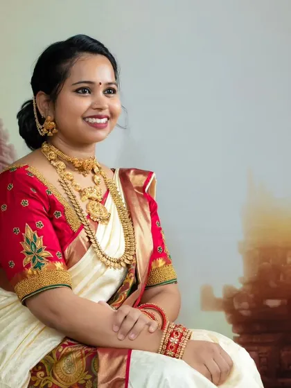 A smiling portrait of a woman in a classic cream and red saree, adorned with traditional temple jewelry.
