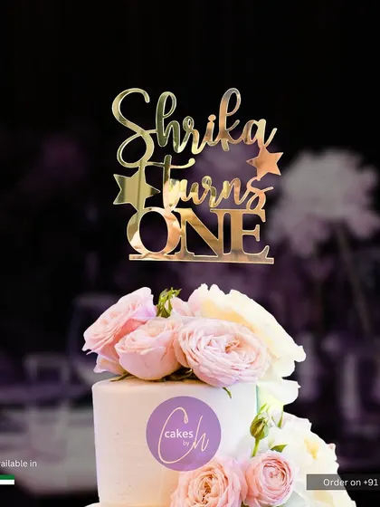 A "Shrika Turns ONE" topper in glossy gold acrylic, shown here on a simple white cake adorned with fresh pink roses. This style combines classic elegance with a personal touch.