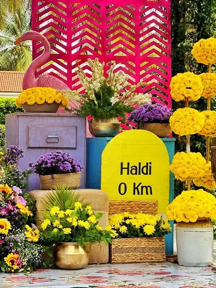 A vibrant and quirky Haldi photo booth, featuring a "Haldi 0 Km" milestone, a pink flamingo, and an explosion of colorful flowers.