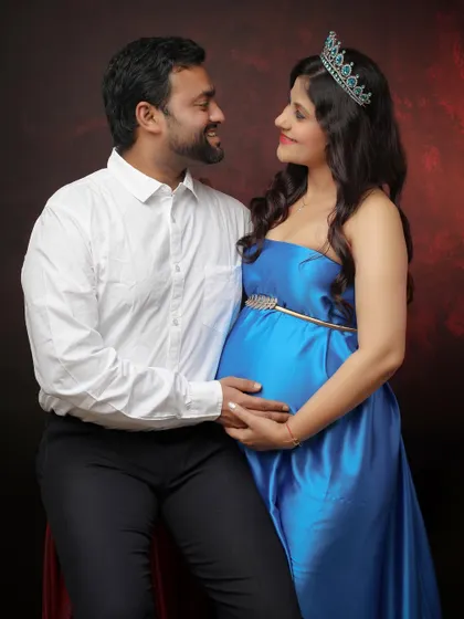 A sweet and classic maternity portrait. The mom-to-be wears a crown and a royal blue gown, sharing a loving look with her partner against a warm, textured backdrop.