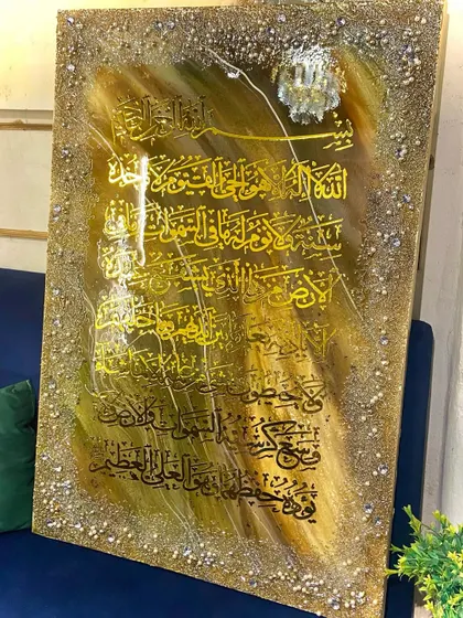 This Ayat al-Kursi piece uses black calligraphy on a gold and brown background, creating a deep, reflective quality. A large crystal accent adds a touch of luxury.