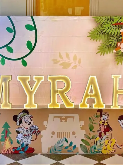 A personalized light-up marquee name sign for "Myrah." The base is decorated with safari-themed illustrations of Mickey and friends, while the top features jungle leaves and a playful monkey.