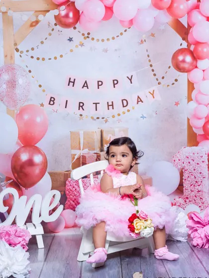 A sweet one-year-old girl in a fluffy pink dress, sitting elegantly in her birthday setup. The pink and white theme with balloons and a "One" sign is perfect for a princess's first birthday.