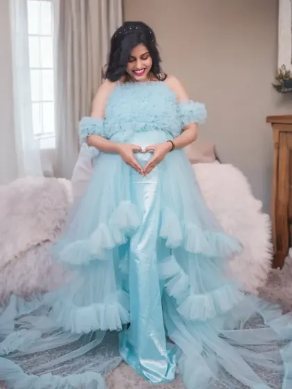 A full-length view of the light blue ruffled gown, showing the tiered layers and sweeping train. The heart gesture over the bump adds a sweet, personal touch to the portrait.