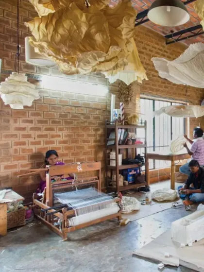 The Oorjaa studio in full swing, with artisans weaving and crafting paper products. The building's honest material palette of brick and concrete provides a robust backdrop for the creative process.