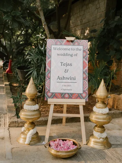 A welcome sign with a traditional Indian textile pattern, flanked by brass lamps, greeting guests to Tejas and Ashwini's wedding.