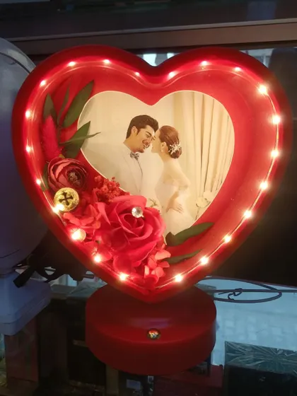 A close-up of our red heart-shaped 3D LED lamp. The photo is surrounded by beautiful artificial roses, and the entire frame lights up to create a romantic glow.