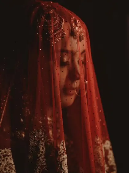 A dramatic, veiled portrait of the bride. The sheer red dupatta with delicate embroidery creates a sense of mystery and timeless romance.