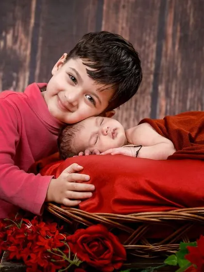 A big brother proudly watches over his sleeping newborn sibling. The rich red tones and rustic wooden background create a warm and loving atmosphere.
