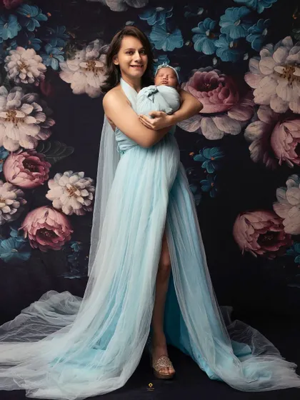 A new mother looking radiant in a flowing blue gown as she holds her swaddled newborn. The floral backdrop adds a touch of artistic elegance to this postpartum portrait.