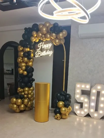 An elegant and modern 50th birthday party decoration. The setup includes a black and gold chrome balloon arch, a 'Happy Birthday' neon sign, and large light-up marquee numbers to mark the milestone.