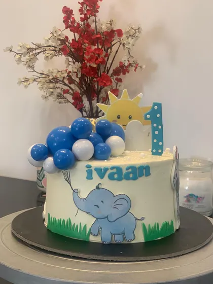 An adorable elephant themed cake for a first birthday. The little elephant is holding a bunch of balloons under a smiling sun.
