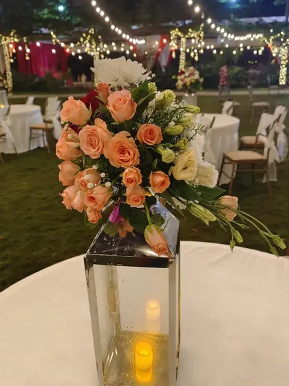 An elegant table centerpiece for an outdoor reception. A lantern with an electric candle is topped with a beautiful arrangement of peach and white roses, adding a soft, romantic glow to each table.