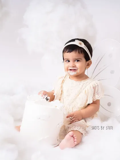 This little angel, surrounded by fluffy clouds, looks so serene and sweet during her fairy-themed cake smash. The all-white setup creates a dreamy and ethereal feel for her first birthday photos.